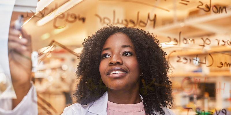 Mastercard Foundation Scholar Charity Bhebhe writes calculations on glass with whiteboard markers while wearing a lab coat. Her ASU research earned her the prestigious Gates Cambridge Scholarship for her doctorate.
